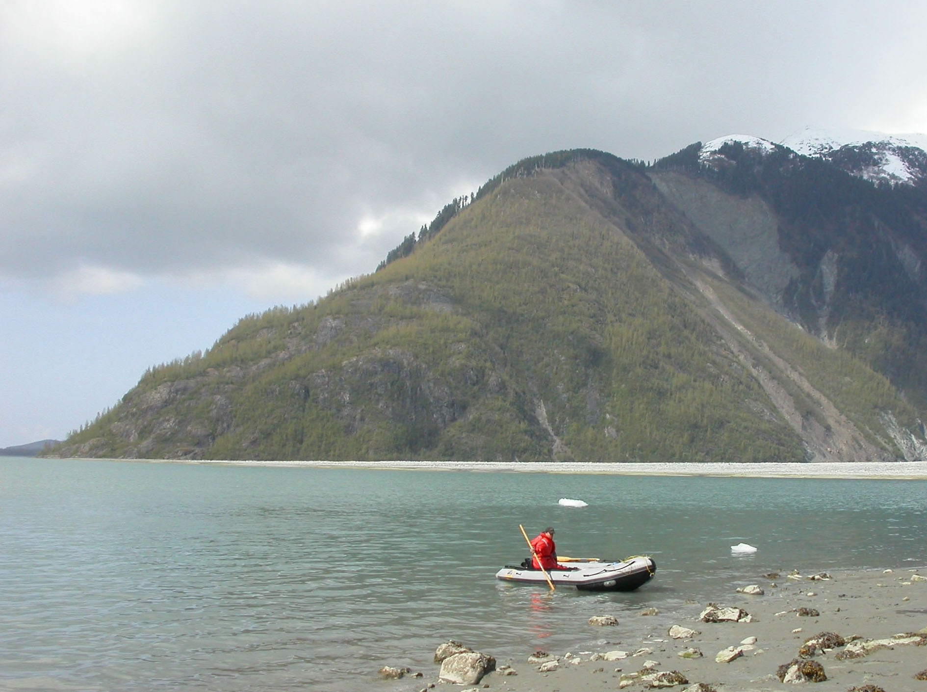 A person wearing a red flotation coat sits in an inflatable boat while bracing an oar in the water near a rocky shoreline. A few ice chunks float nearby. In the background, a steep, snow-topped mountain rises from the waterline. 