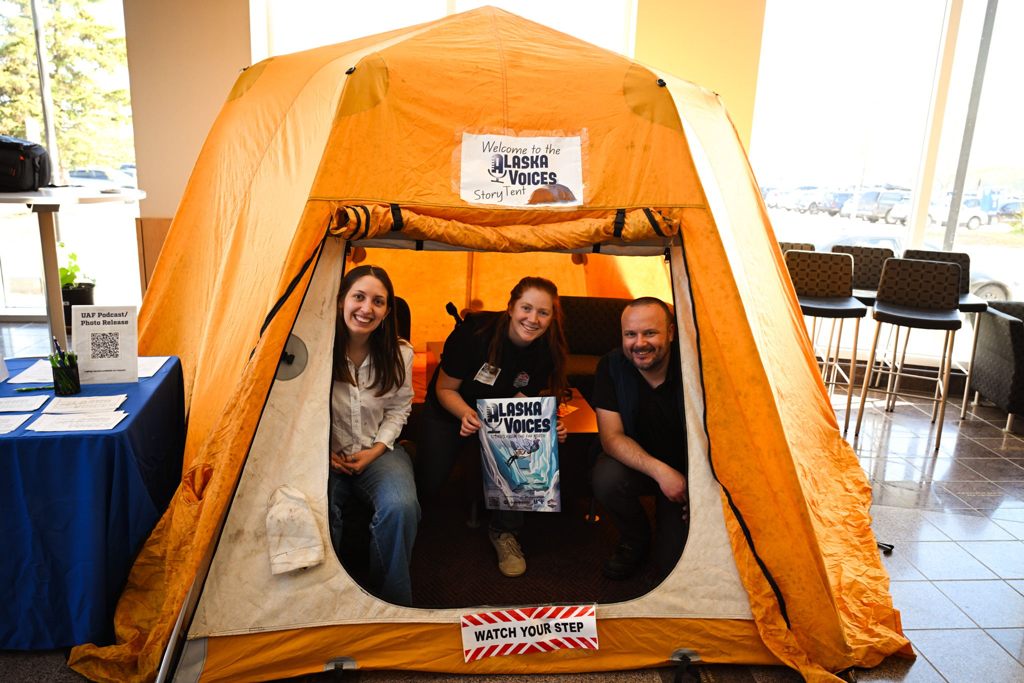 Three people smile insides a yellow tent, inside a building. They hold a poster that reads Alaska Voices. The tent is labeled the Alaska Voices Story Telling Tent.