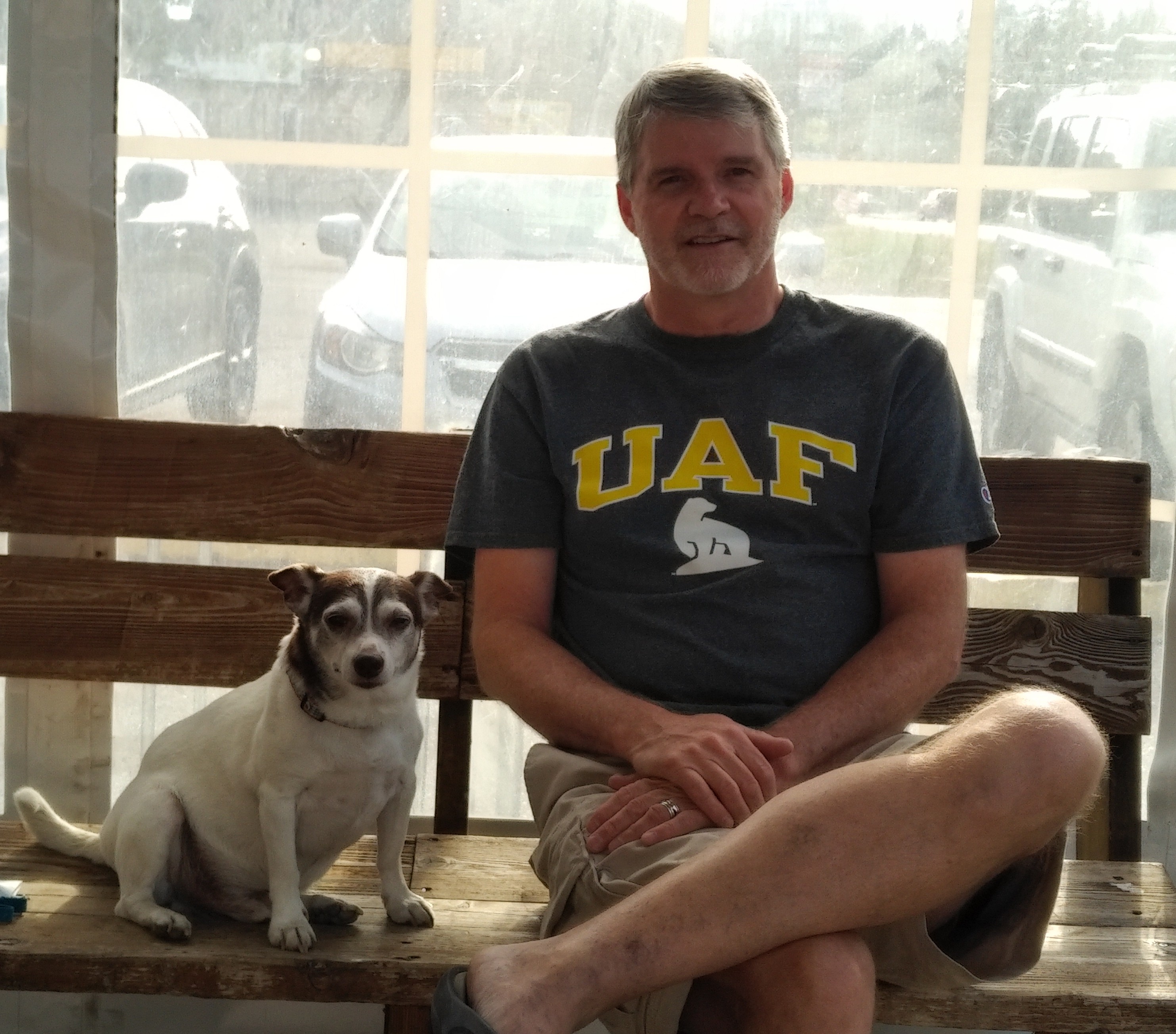 a little dog to the left and a guy on the right sitting on a wooden bench