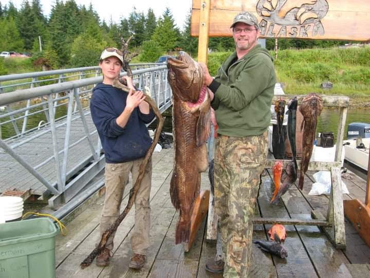 A women standing to the left holding large drift wood as comparison to the large Lingcod fish a man is holding.