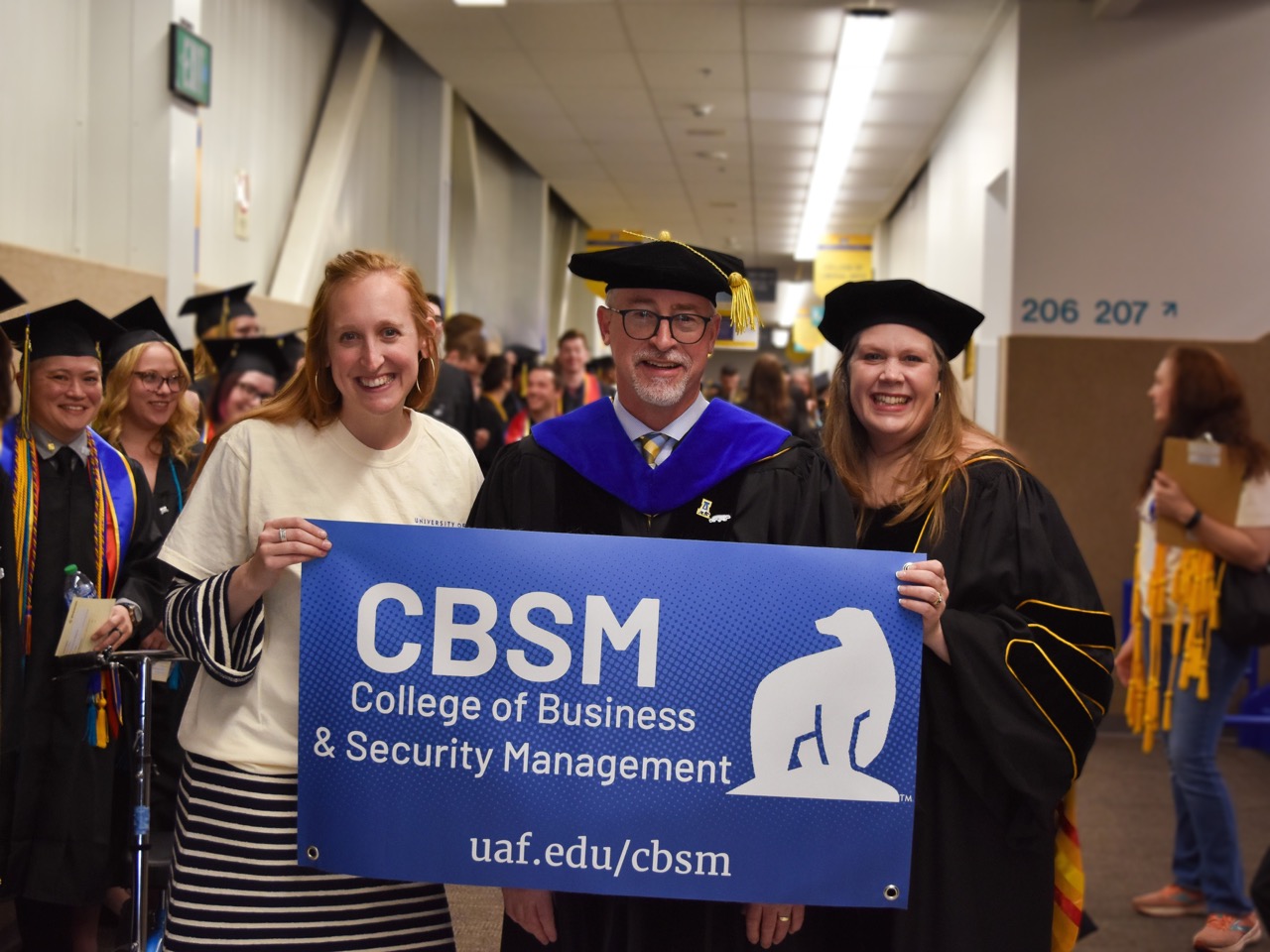 a man stands between two women in graduation regalia holding a blue banner that reads CBSM College of Business and Security Management uaf.edu/cbsm