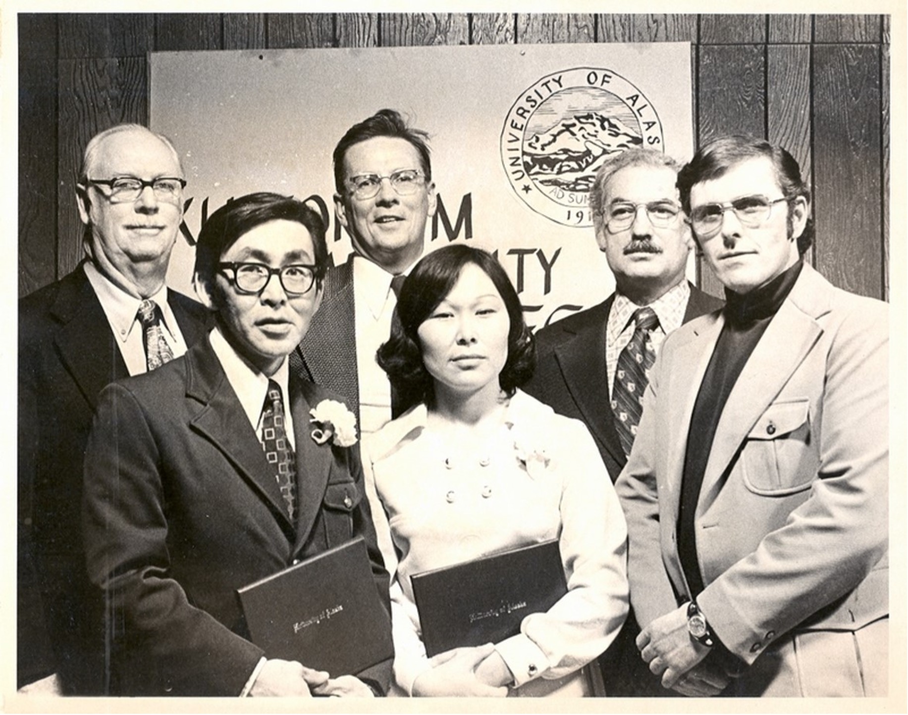 Black and white photo from 1975 of a group of people. A man and woman in the front row hold diplomas, with four men standing in a row behind them. The group is in front of a partially obscured banner showing the University of Alaska logo.