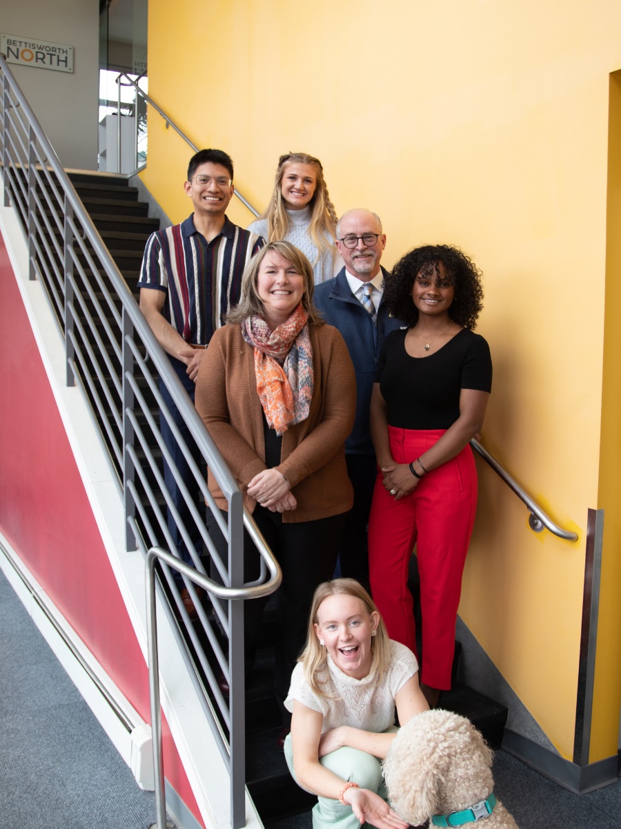 Tracy Vanairsdale stands on staircase with CBSM Dean Cameron Carlson, three students and her dog, Copper.