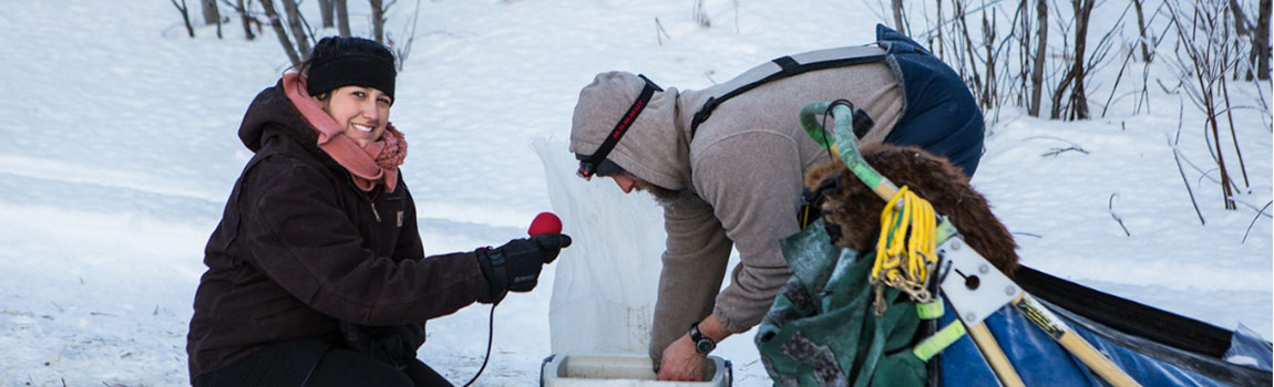 A kneeling journalism student smiles at the camera while holding a microphone to interview a musher, who is bending to reach into a small cooler near his dog sled