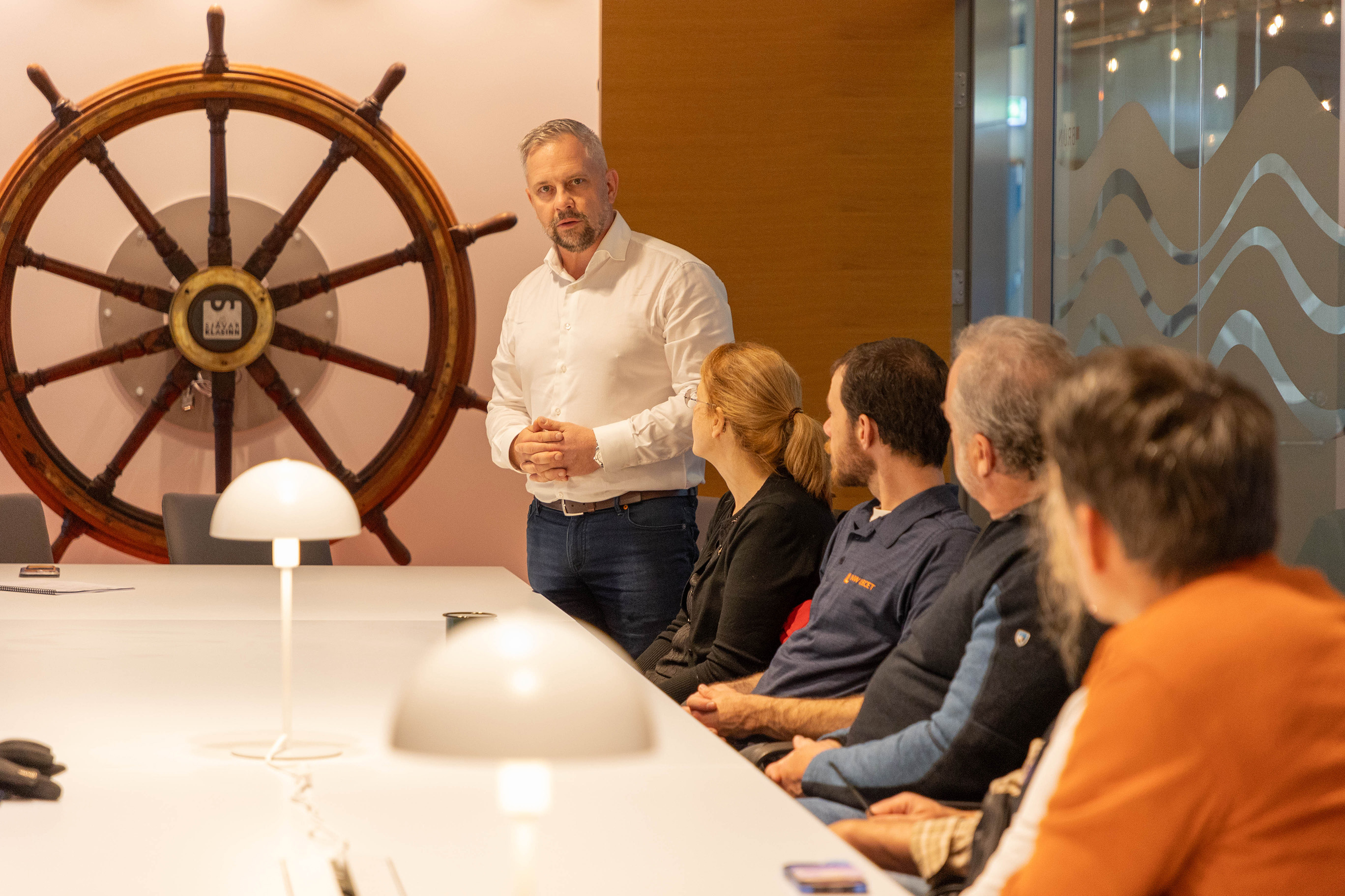 A person standing is answering questions to a group of people who are sitting around a table.