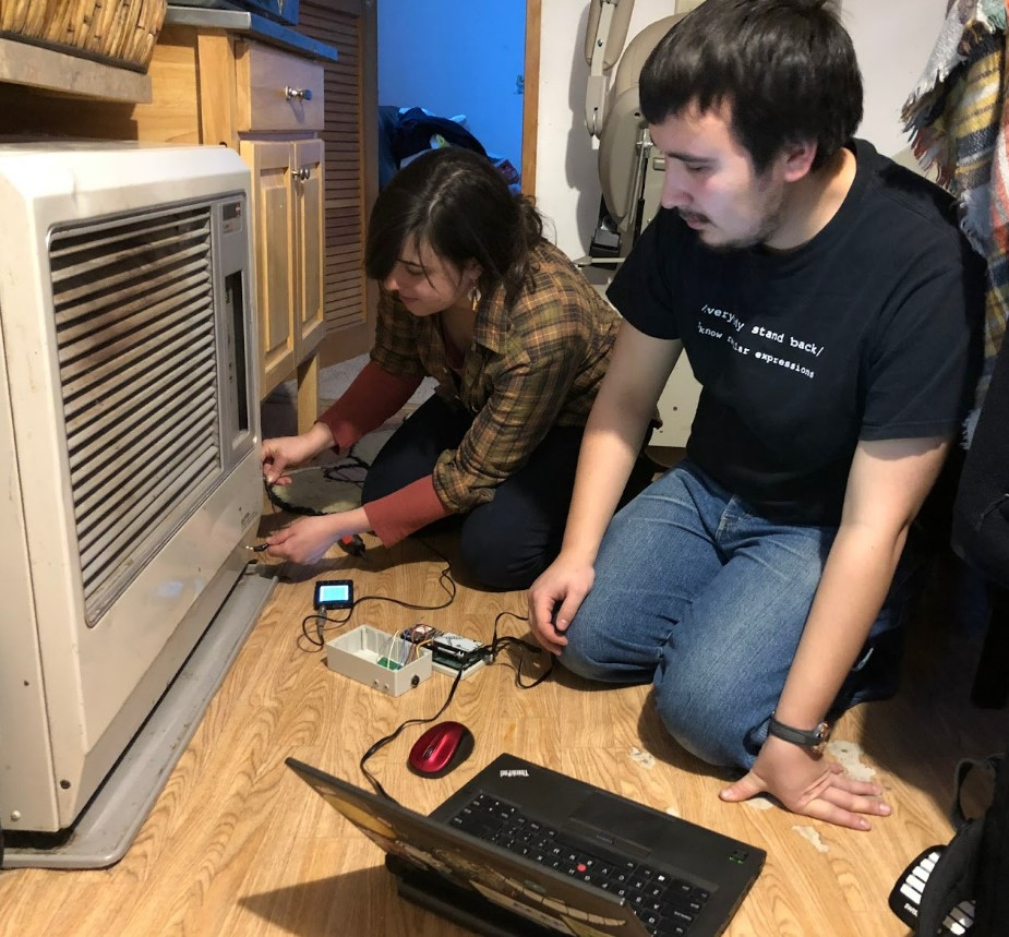 Two people on the floor installing a fuel consumption monitoring device on a stove.