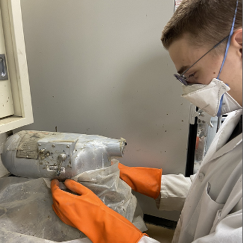 A person wearing a protective mask and gloves handles a bag of ash-like substance in a lab.
