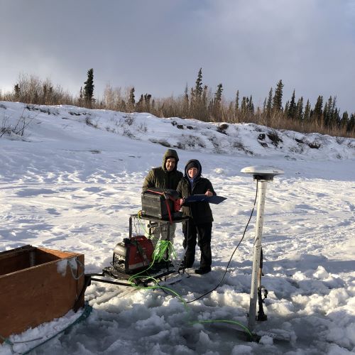 Two researchers stand in the winter sun on the frozen Yukon river.