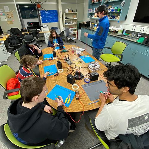 A group of people sitting at a desk working on their electrical soldering project while a person standing and giving them instructions.