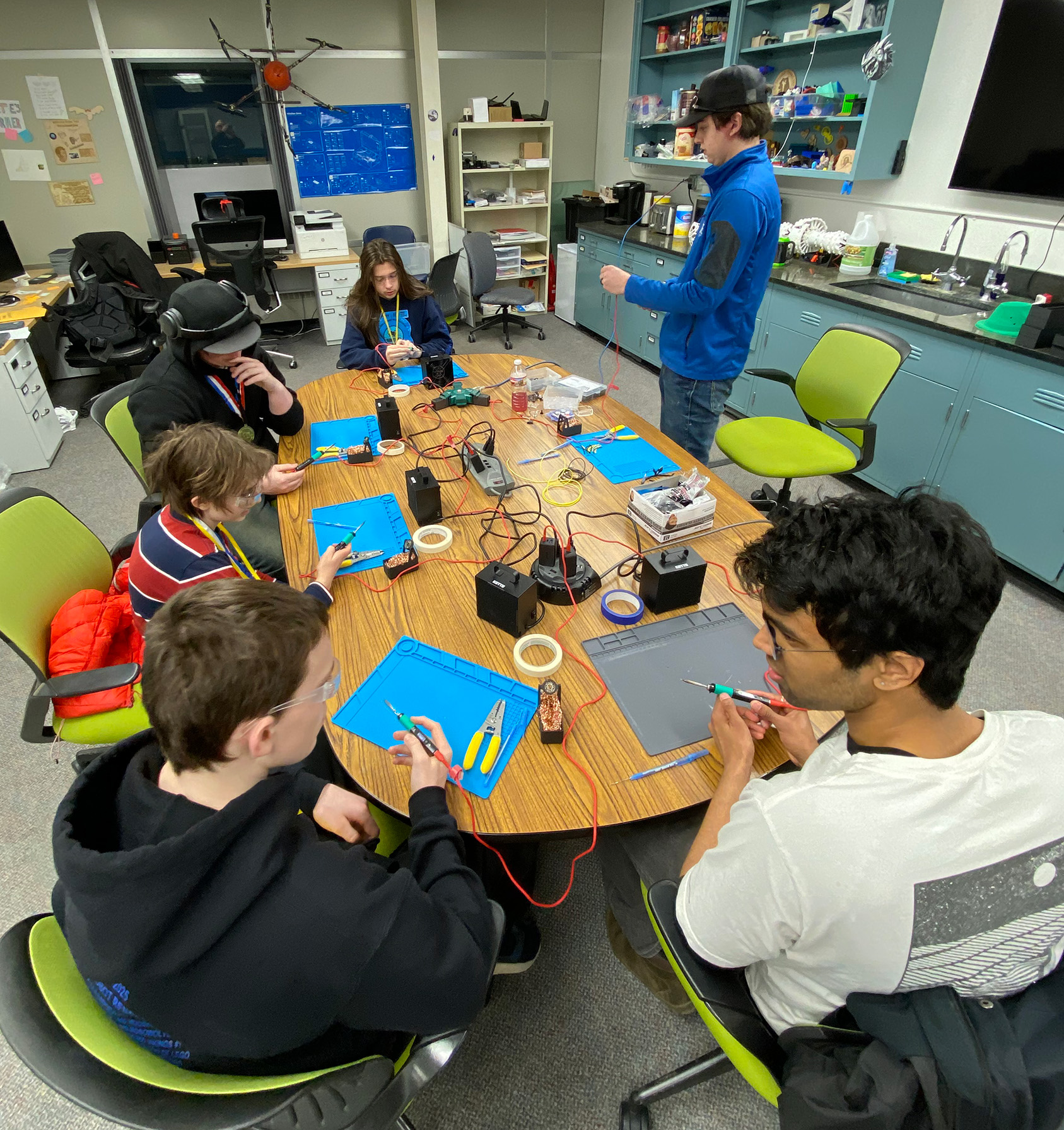 A group of people sitting at a desk working on their electrical soldering project while a person standing and giving them instructions.