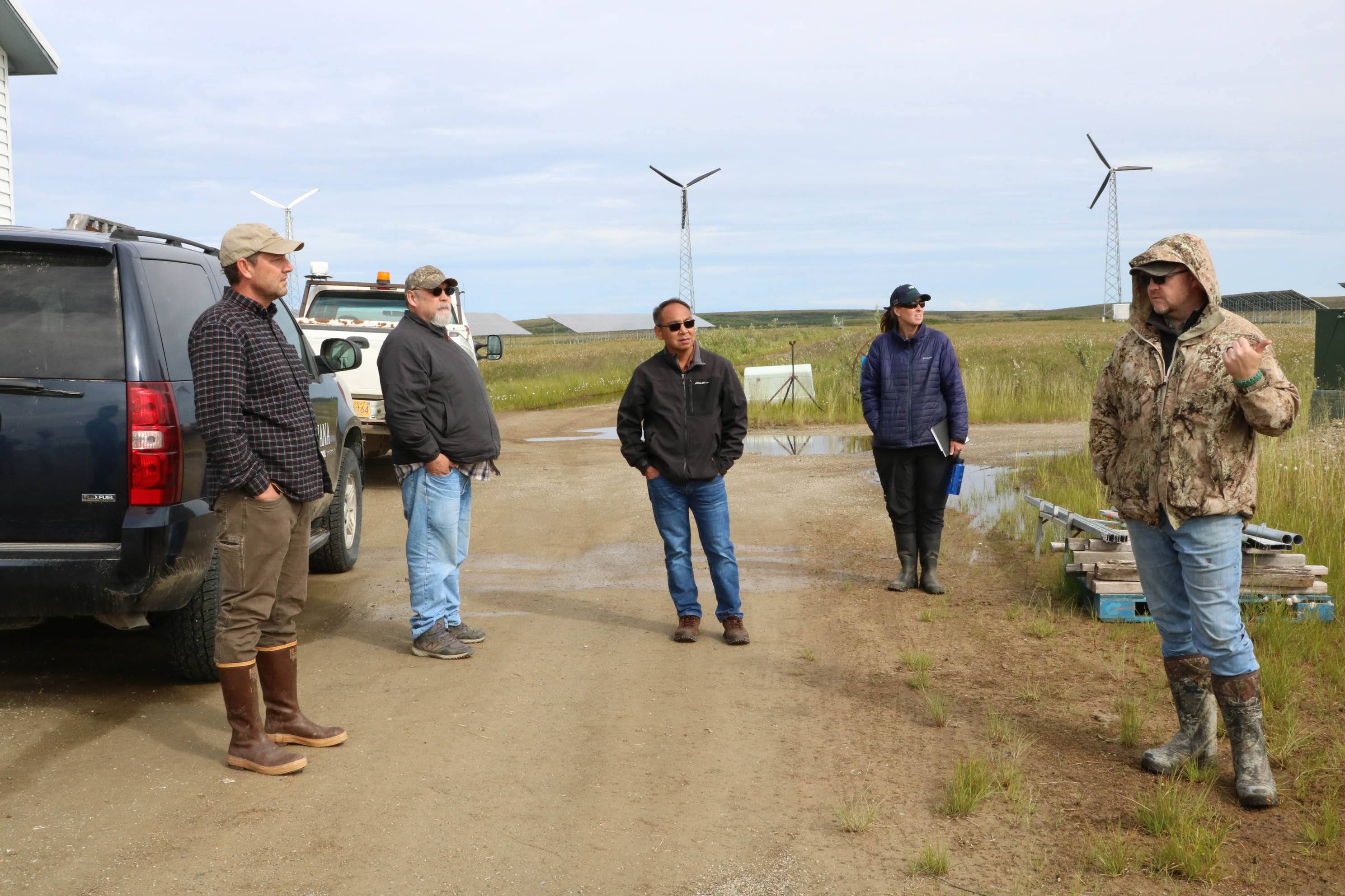 People stand on a dirt road where wind turbines and solar panels visible in the background