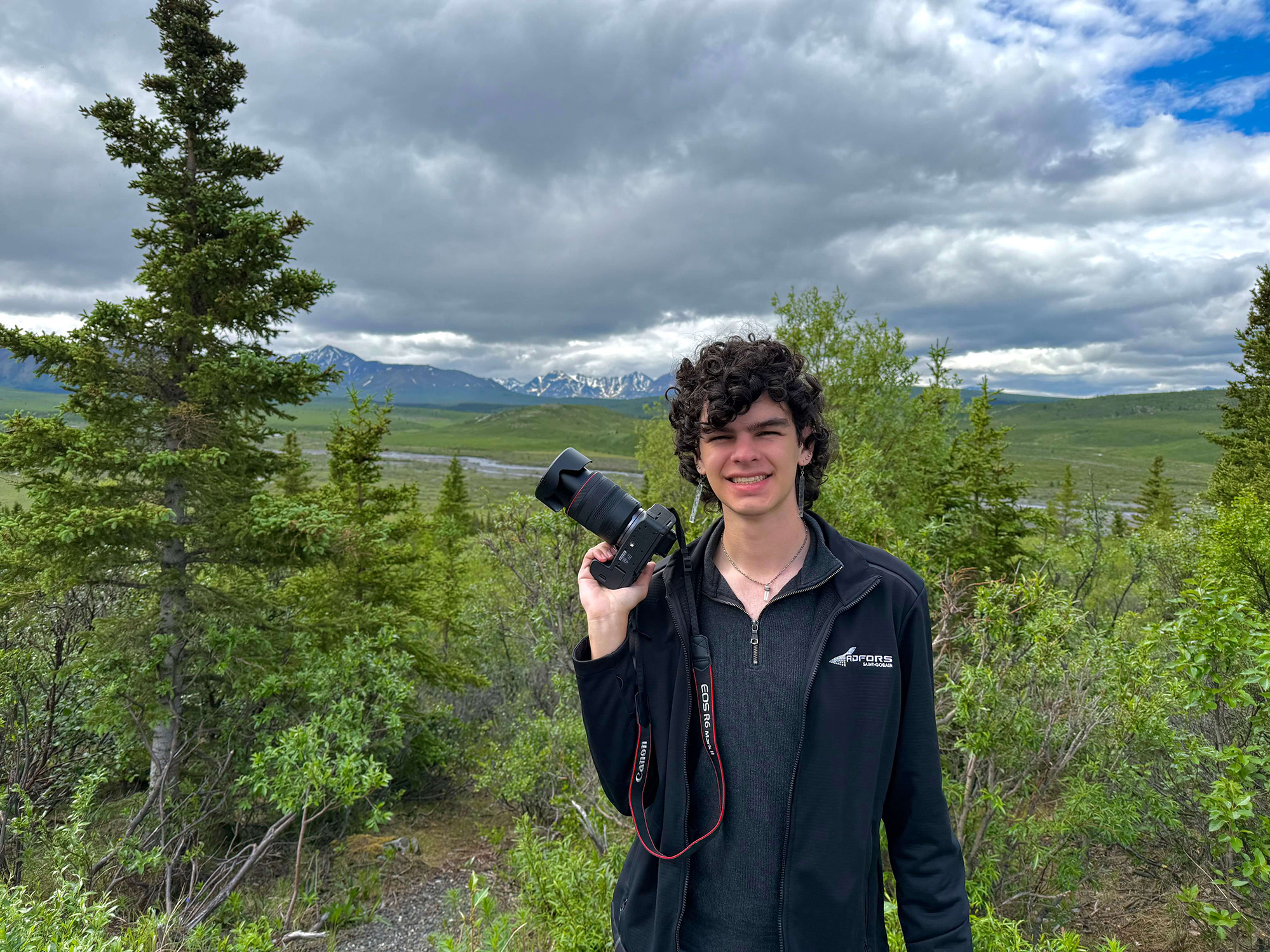 A person holding a camera stands in a field with trees and sky in the background