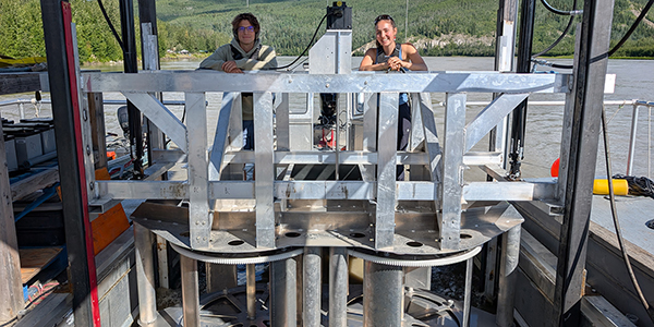 Two people standing behind a turbine on a ship on a river.