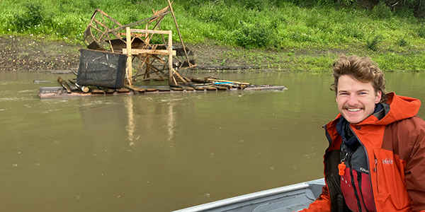 A person is sitting in a boat on a river with a traditional fish wheel in the background.