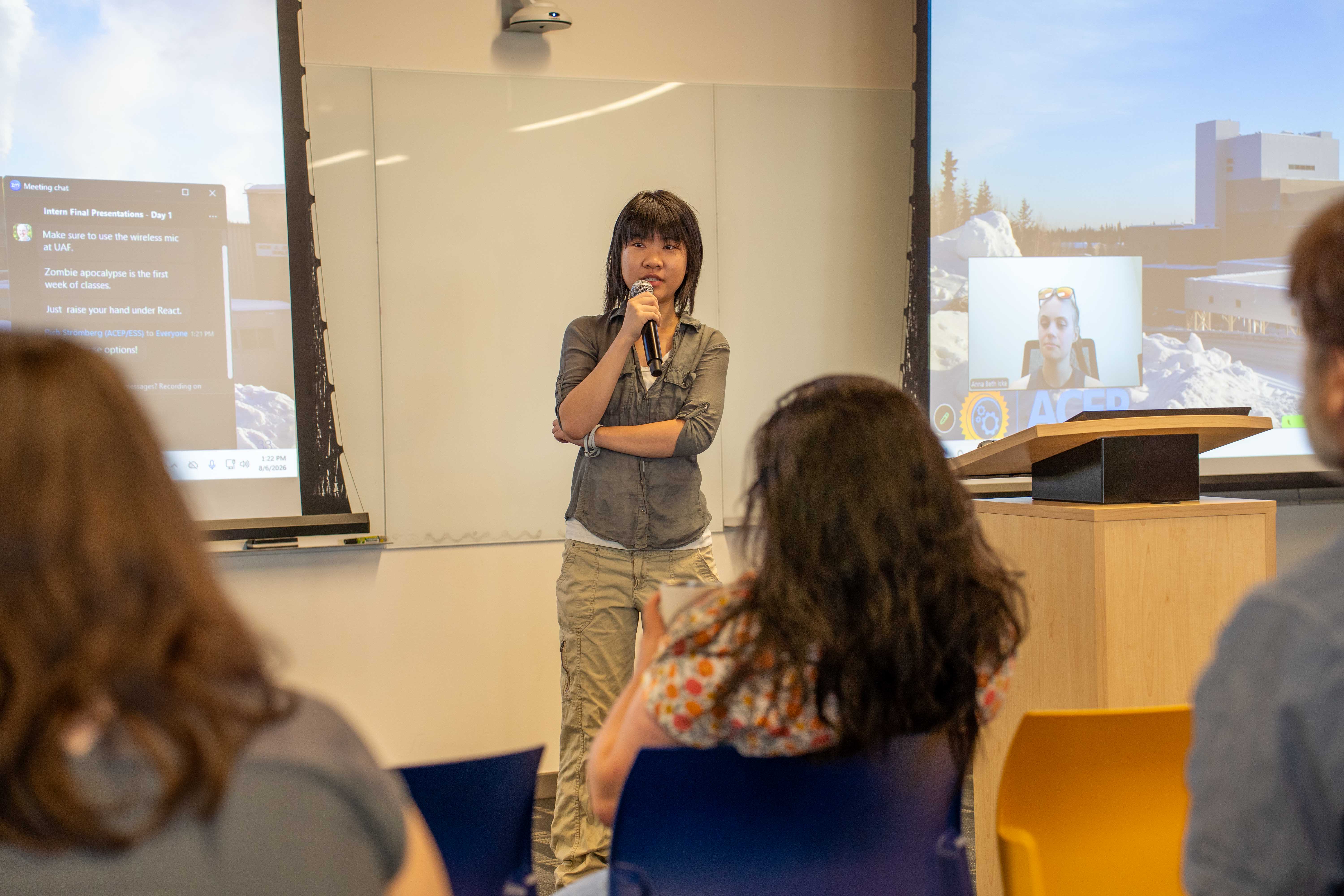 A person with a mic giving a presentation with two screens behind her