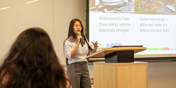 A person giving a presentation with a microphone in hand in front of a screen.