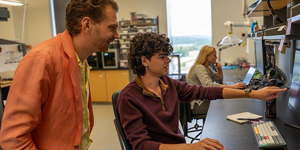 A person is pointing at a computer screen at a desk while the other person is standing looking at the screen.
