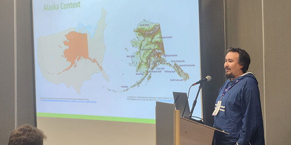 A person is giving a presentation with a screen showing Alaska maps on his right.