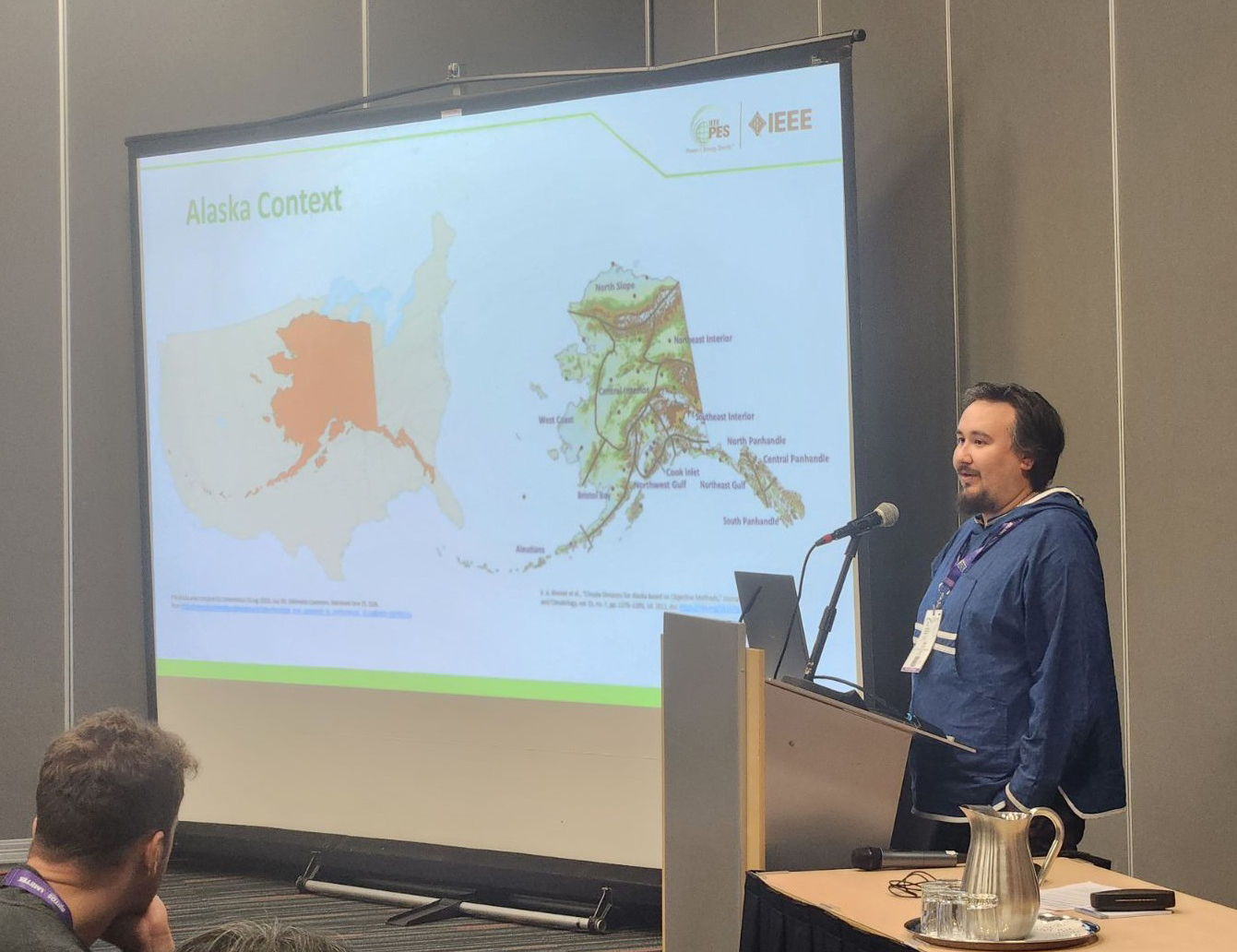 A person is giving a presentation with a screen showing Alaska maps on his right.