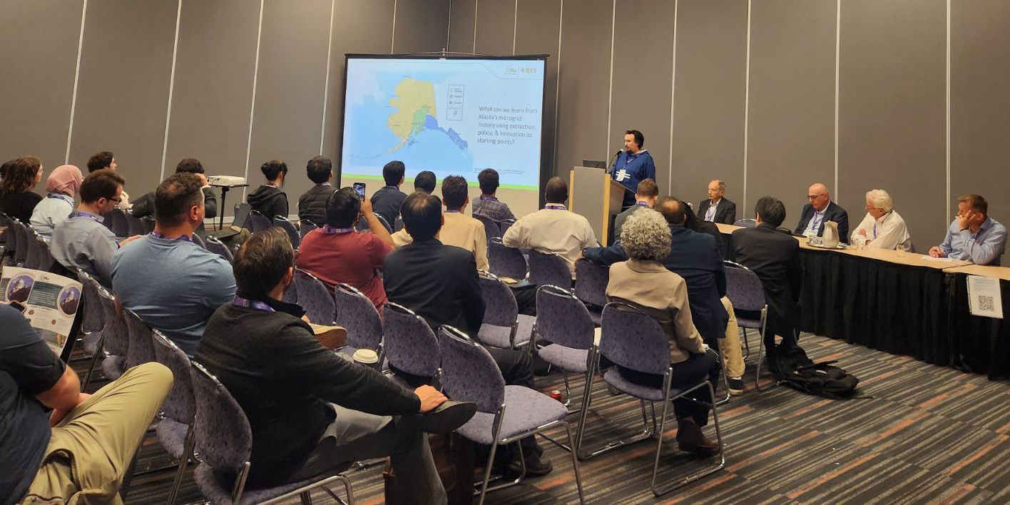 A person is giving a presentation to a group full of people. Four other panelists sit at the table on his left.