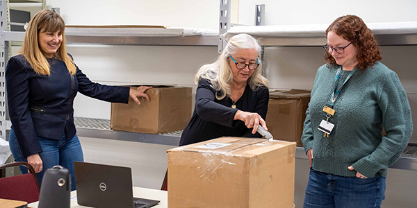 A person is opening a box placed on a desk, while two people look on.
