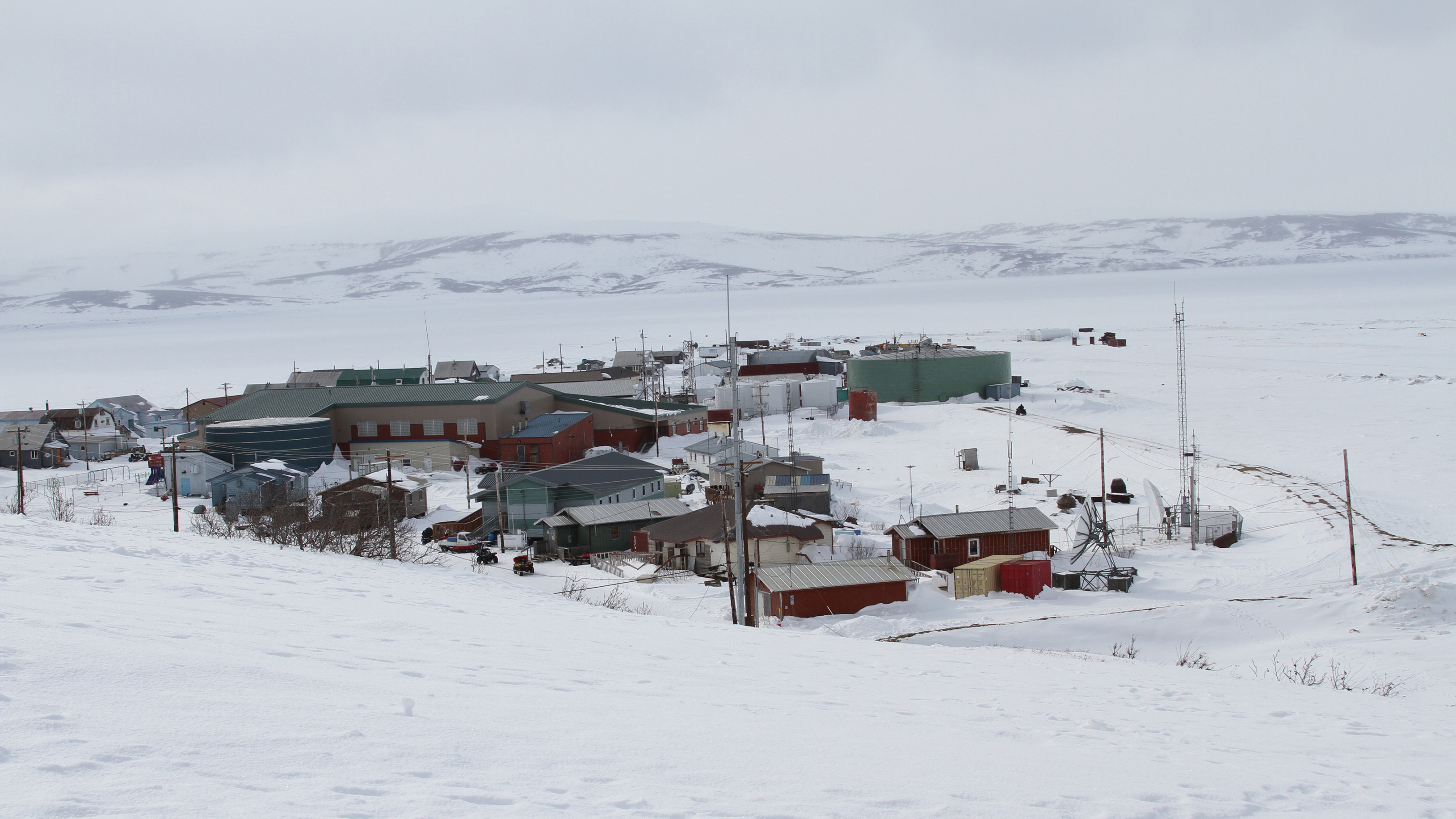 Houses and other buildings in a small Alaska village in winter. Mountains are seen in the far distance. 