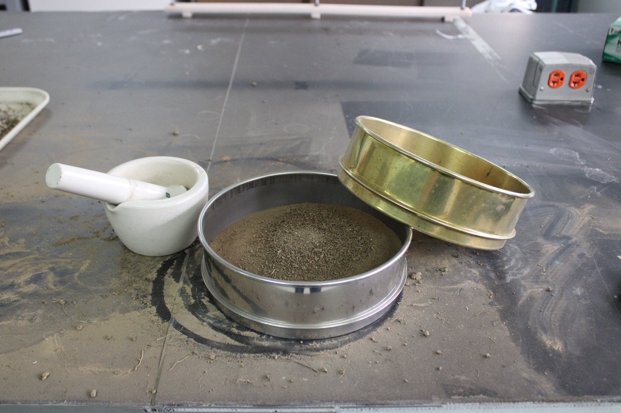A white motar and pestle sits next to a silver soil sifter filled with soil. These are tools used to process soils