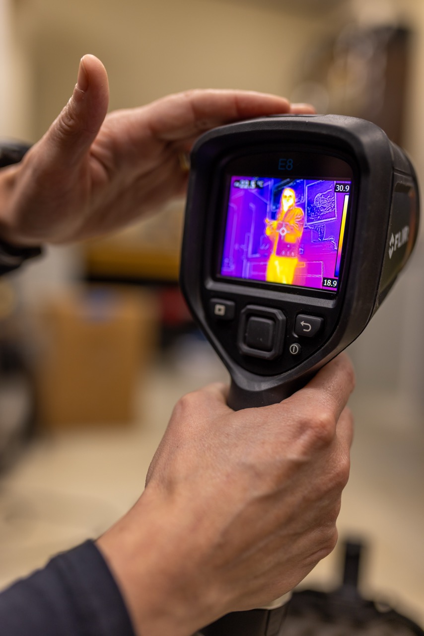 A handheld camera screen shows the thermal heat map in yellows and oranges of a person standing