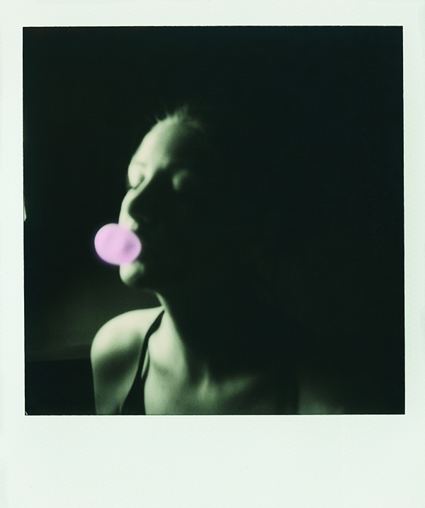 A Polaroid portrait of a woman blowing a bubble with gum. The portait is in black and white and the bum is hand colored pink