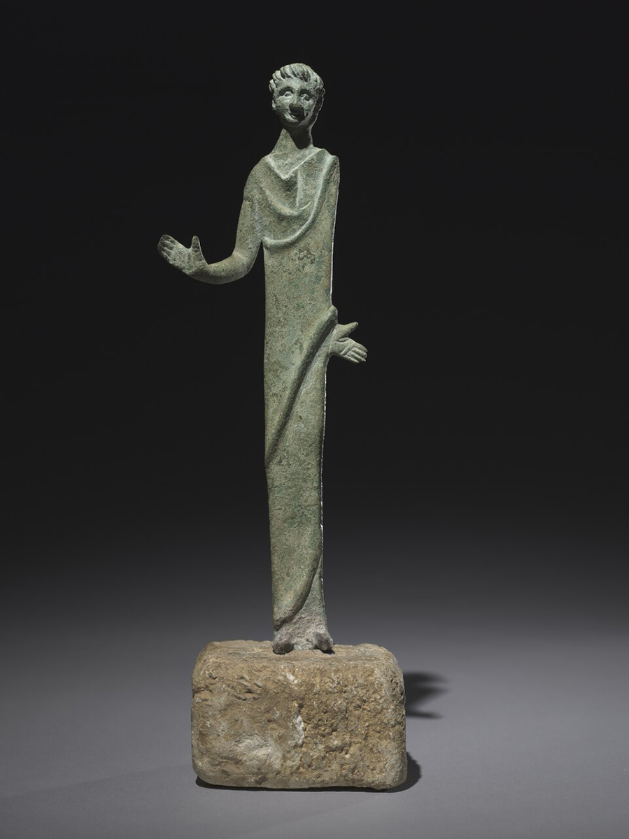 Small bronze Etruscan votive figure of a standing man with elongated proportions and simplified features. The figure wears a long draped garment and gestures outward with both hands. Its stylized face and rigid posture reflect the abstract, expressive qualities common in Etruscan bronze sculpture. The sculpture stands on a rough rectangular stone base against a dark background.