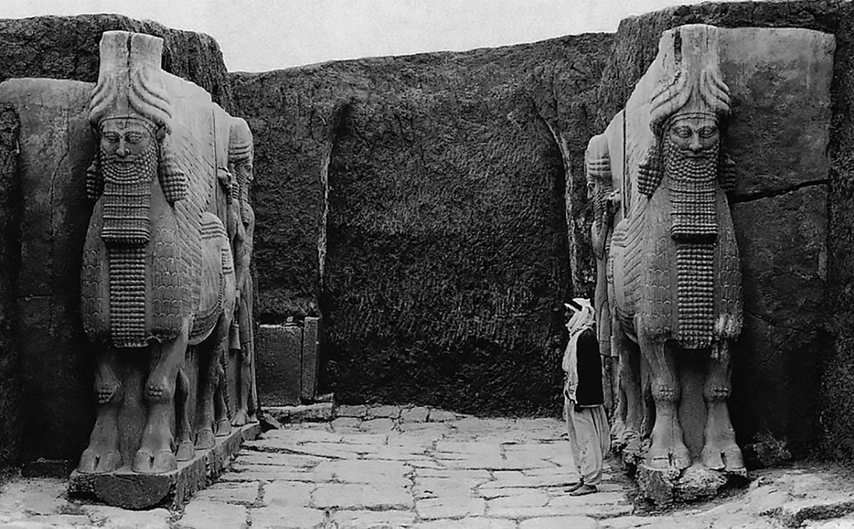 Colossal Assyrian lamassu sculpture combining the body of a winged bull or lion with a human head wearing a royal headdress. The guardian figure stands in a powerful stance with detailed musculature, layered wings and a long curled beard. Carved in relief from stone, the lamassu originally flanked the gateway of the citadel of Sargon II, serving as both protector and symbol of royal authority.