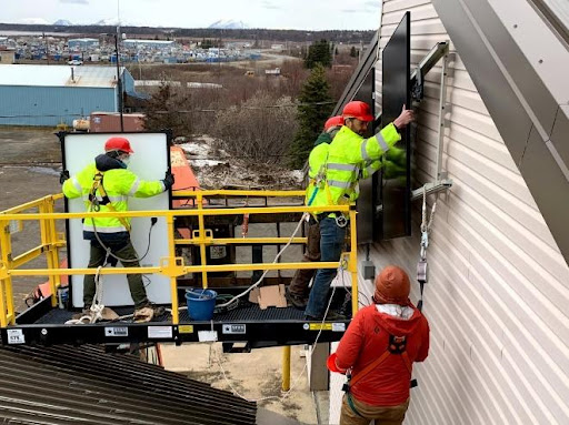Students standing on lift work together to attach a solar panel to the side of a building. 