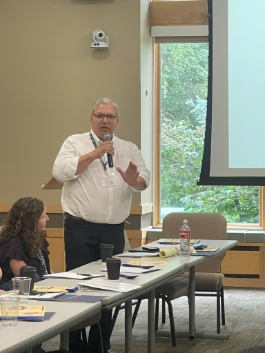 A person stands behind a table with a microphone and addresses the room.
