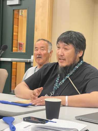 A seated man speaks behind a table while someone listens behind him. 