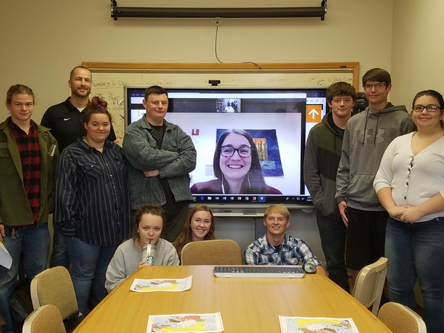A group of people stand around a video screen with a picture of a woman videoconferencing in on it.