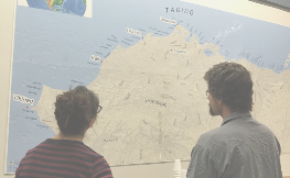 Two people stand with their backs to us, looking at a map of Alaska