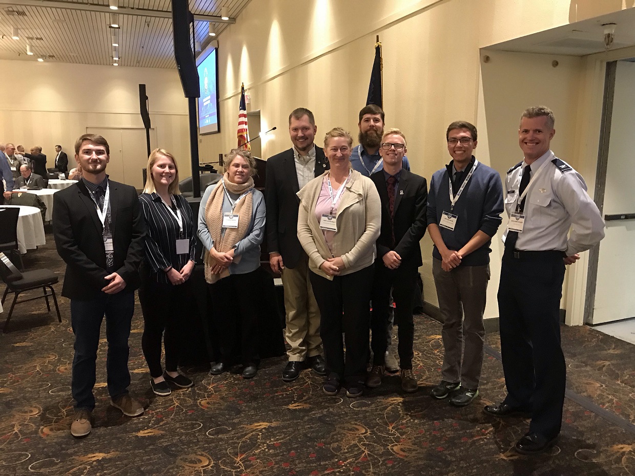 Nine people wearing badges stand together in a conference meeting room. 