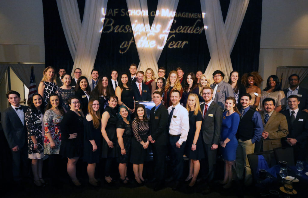large group of students dressed up in dresses and suits and ties pose together in front of the UAF School of Management - Business Leader of the Year stage