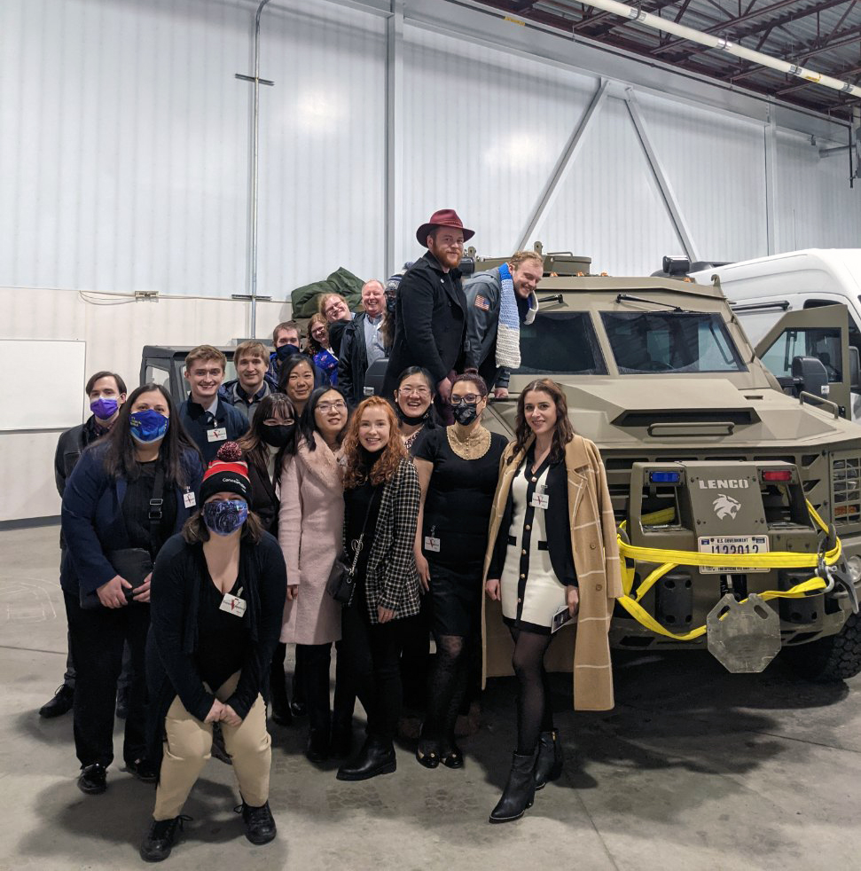 large group of about twenty students pose in front of army vehicle in a garage
