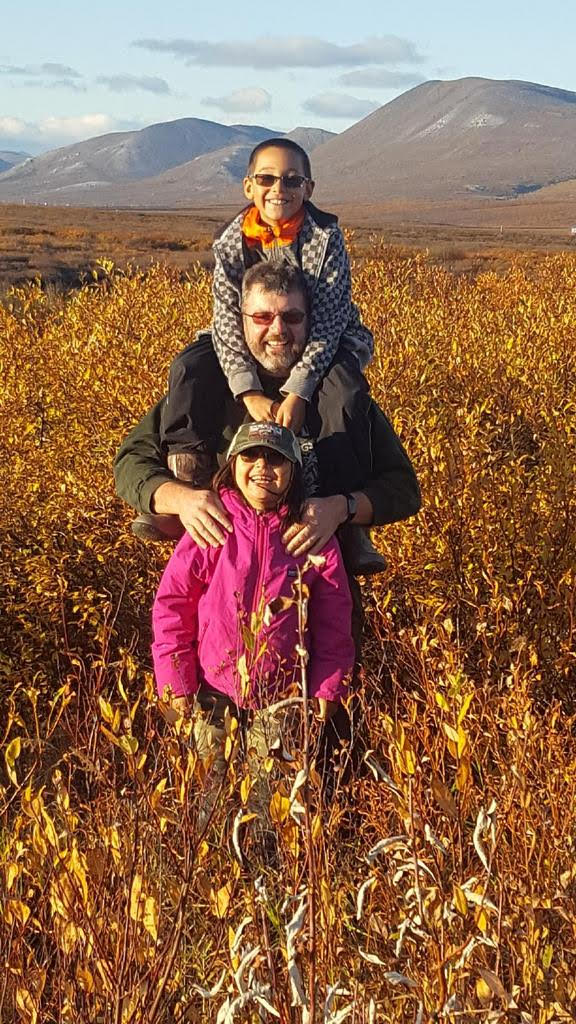 Father stands in field with son on his back, and daughter in front. Fall time, mountains in background
