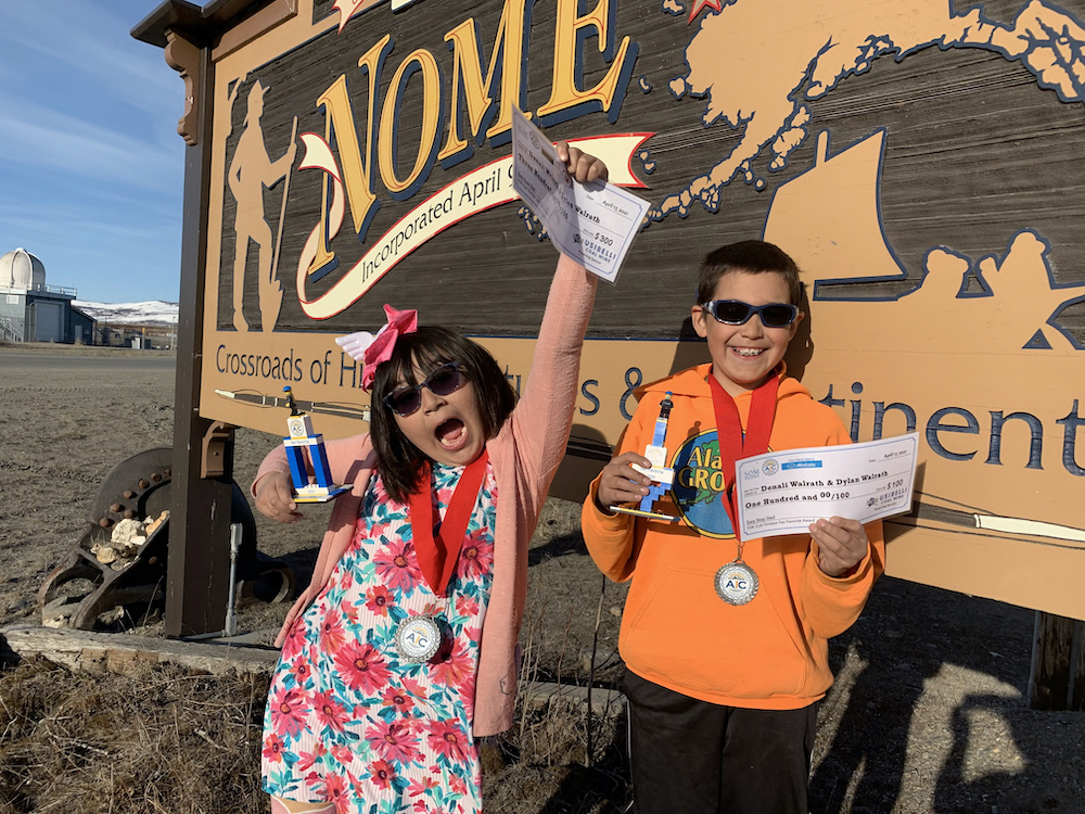 young sister and brother hold up trophies and checks in front of the Welcome to Nome sign in Nome Alaska