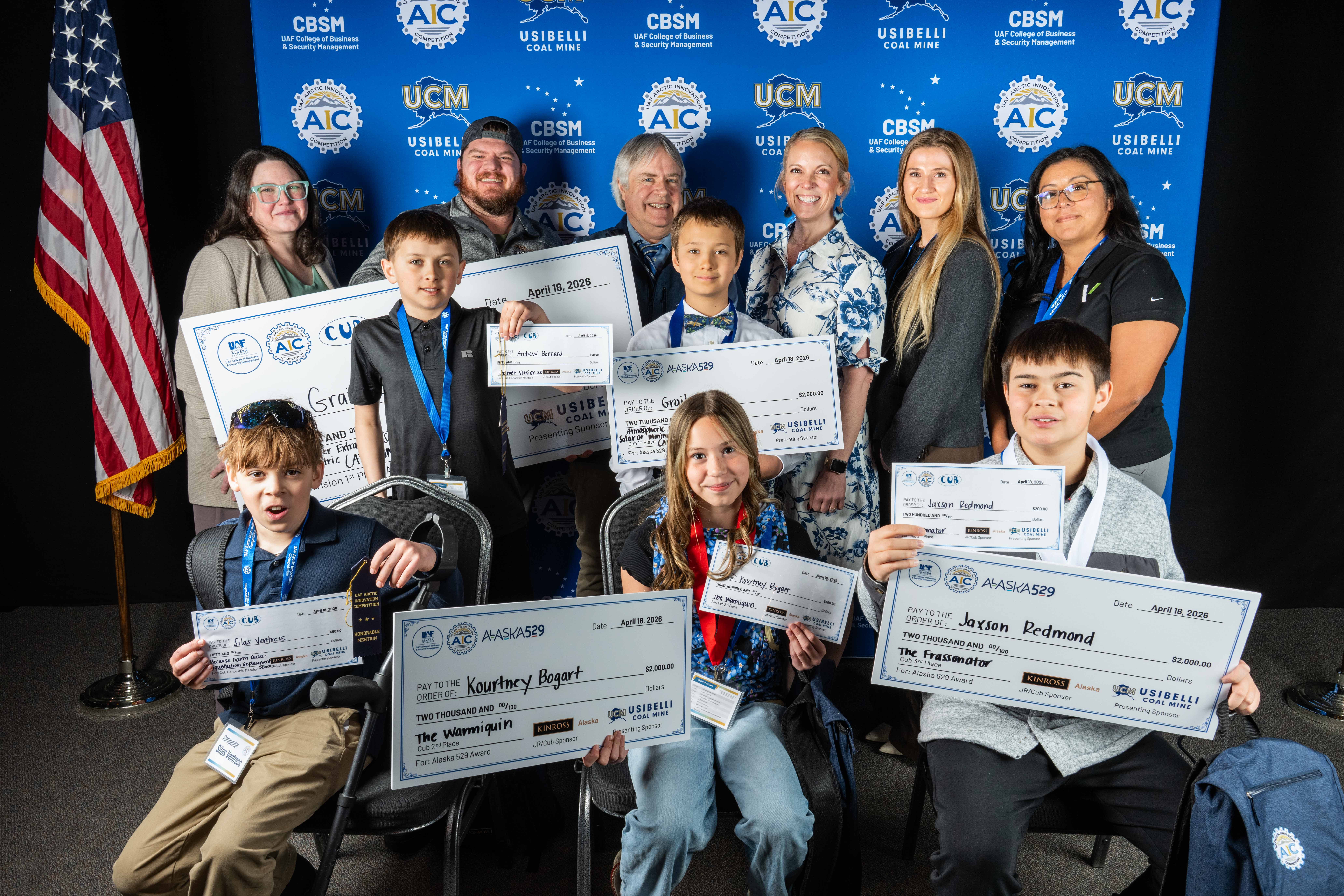 two boys hold up giant checks in front of blue backdrop that reads CBSM College of Business and Security Management and Usibelli Coal Mine