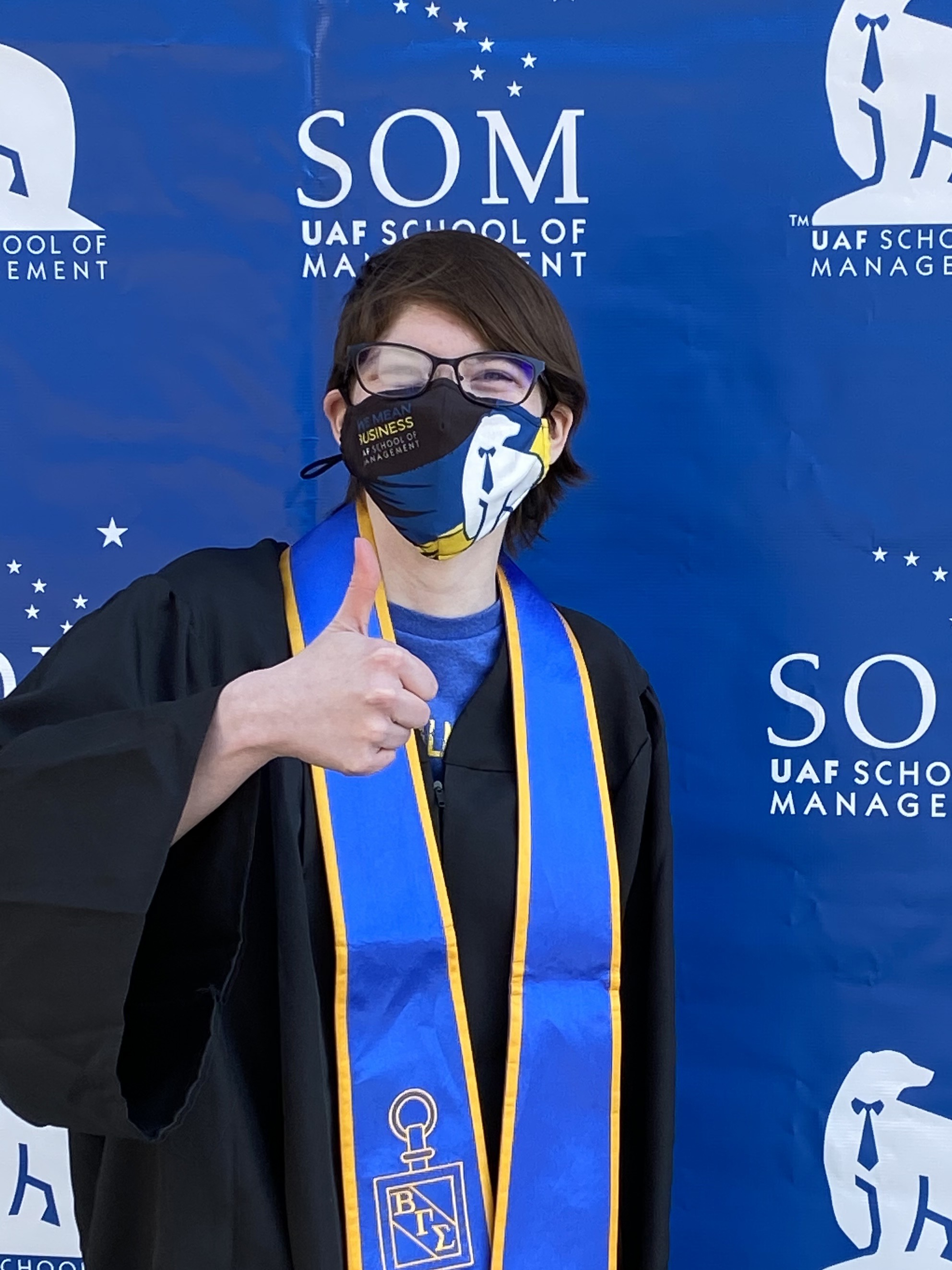 college graduate gives thumbs up wearing regalia and a mask