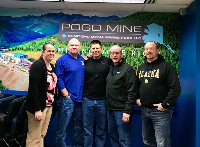 four men and one woman pose together in front of wall with Pogo Mine logo