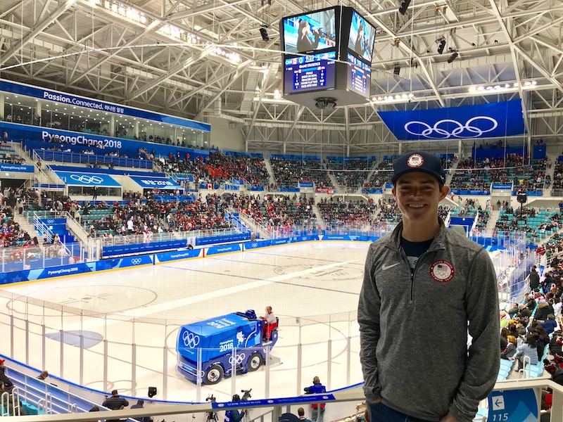 man smiles in sport jacket and ball cap in front of ice arena