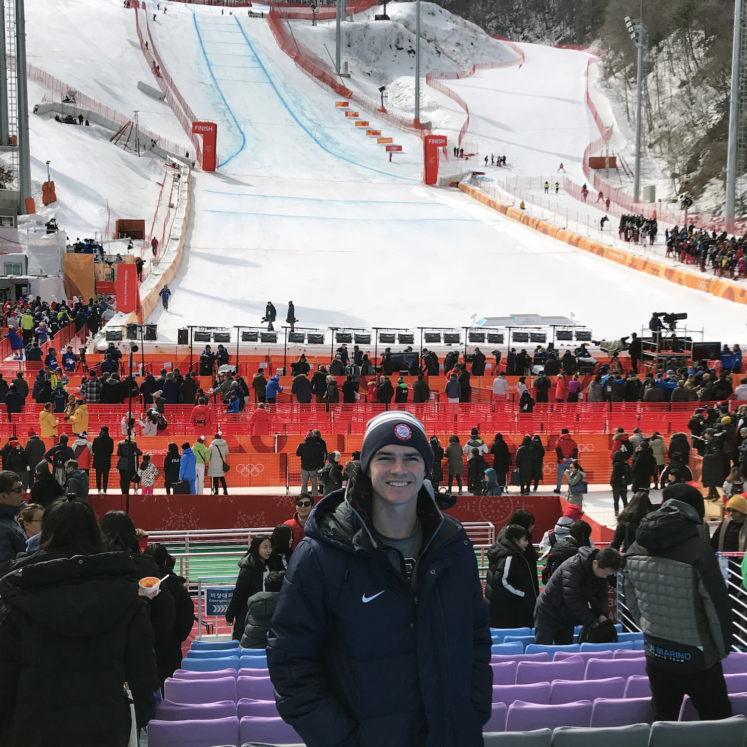 man smiles wearing winter jacket and beanie outside in front of ski race hill