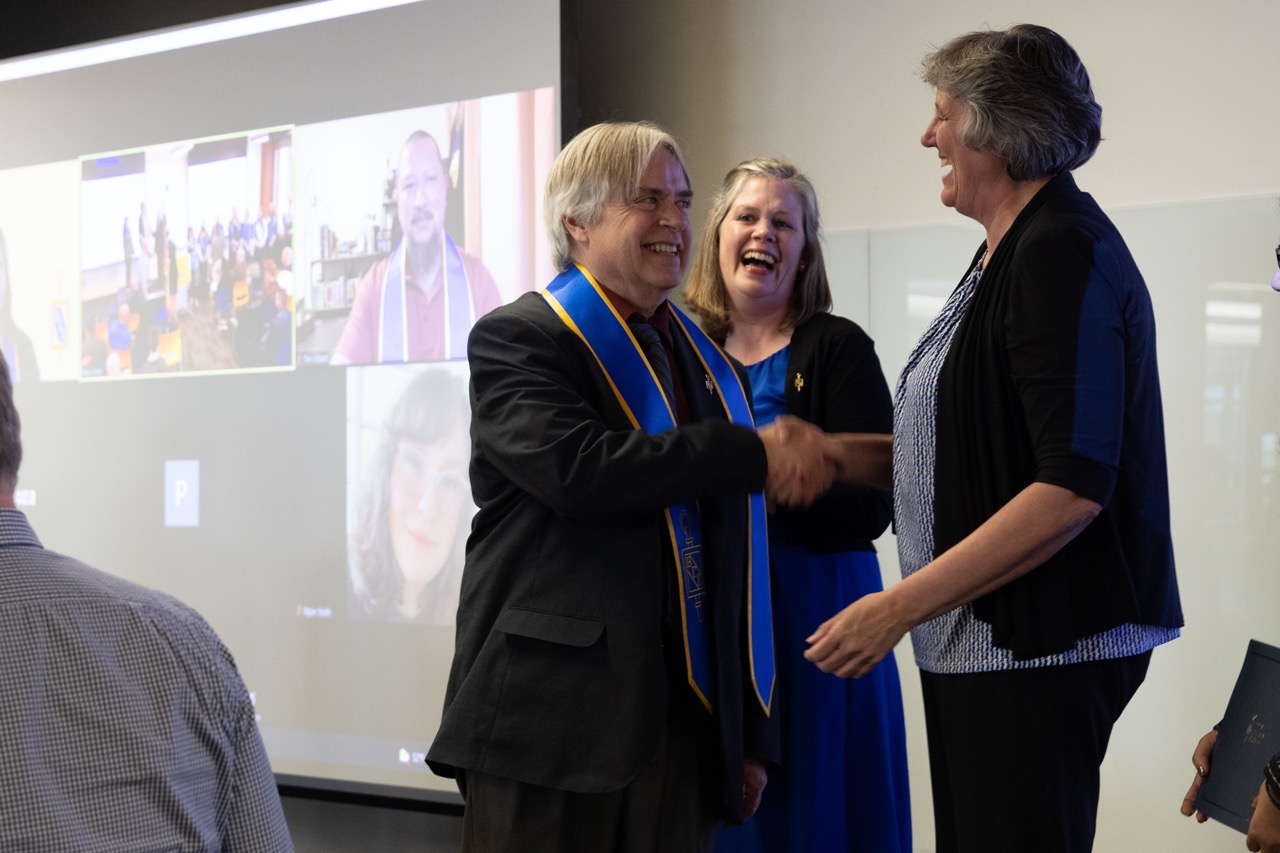 man shakes hand with woman while wearing blue and gold sash. woman in background laughs happily