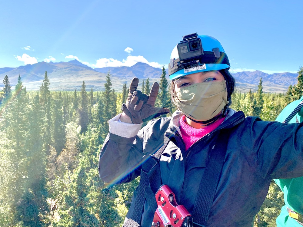 woman in zipline gear takes a selfie with mountains