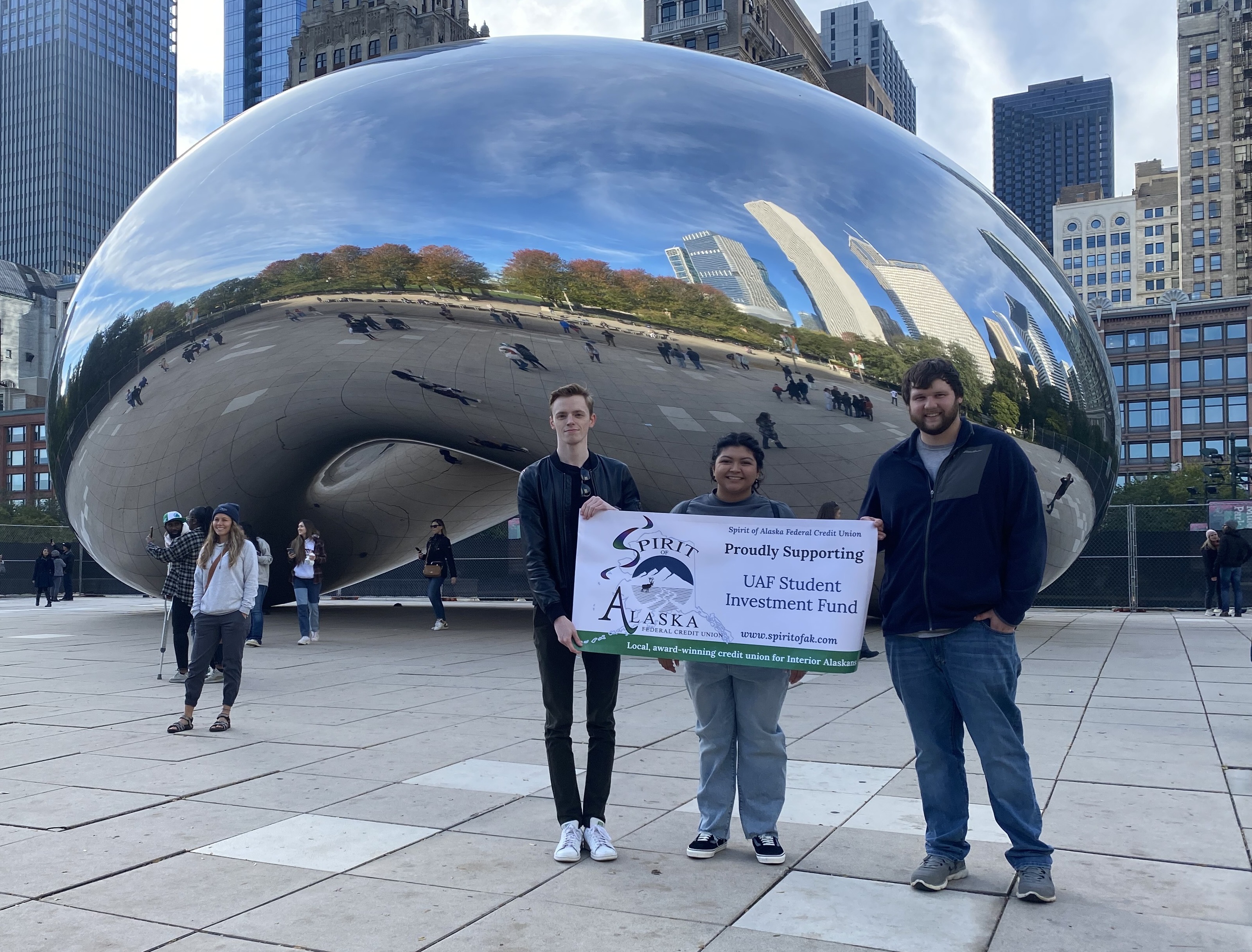 three students hold Spirit of Alaska banner in front of "The Bean", a silver sculpture in Chicago