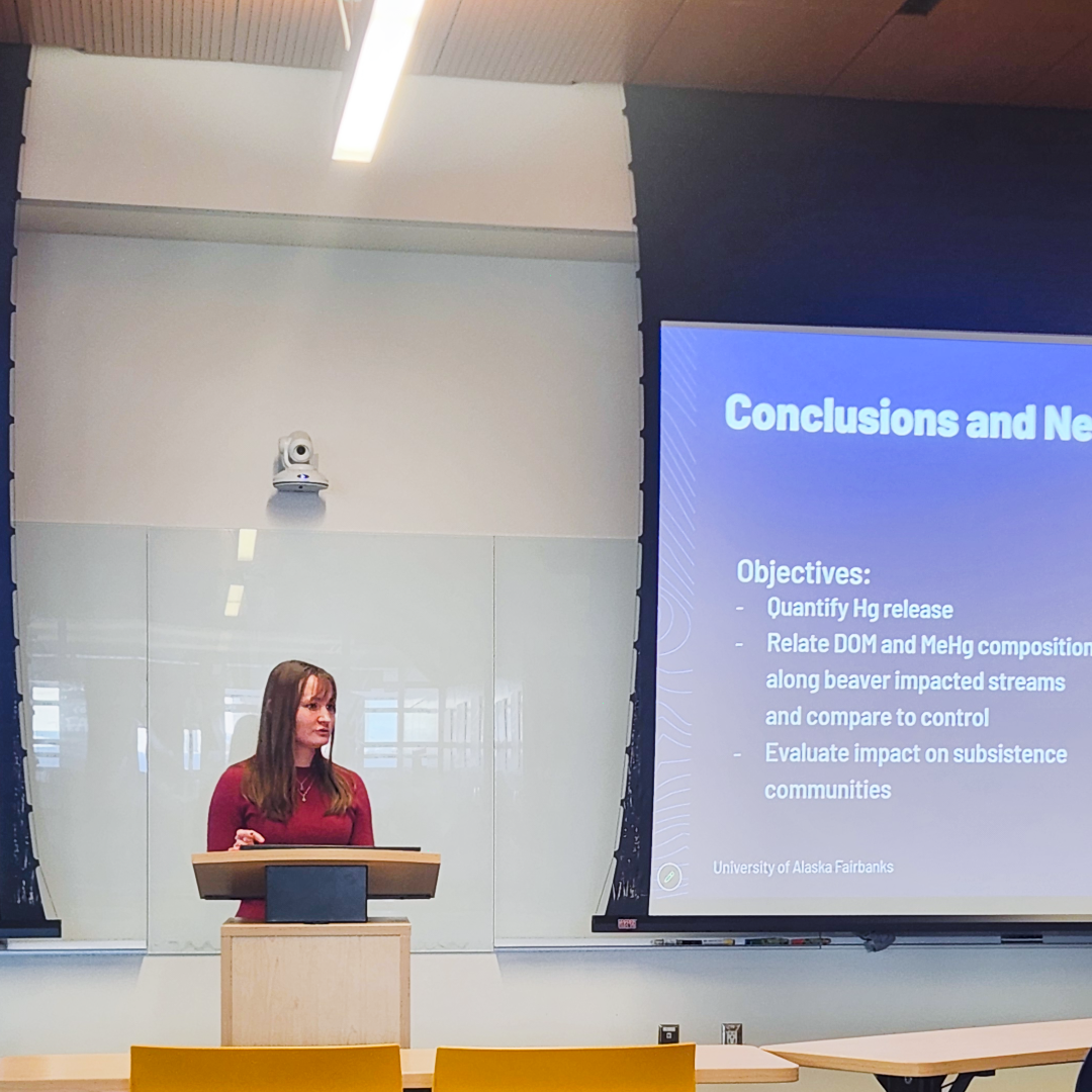 A scientist presents her research at a podium with dual project screens behind her in a classroom.